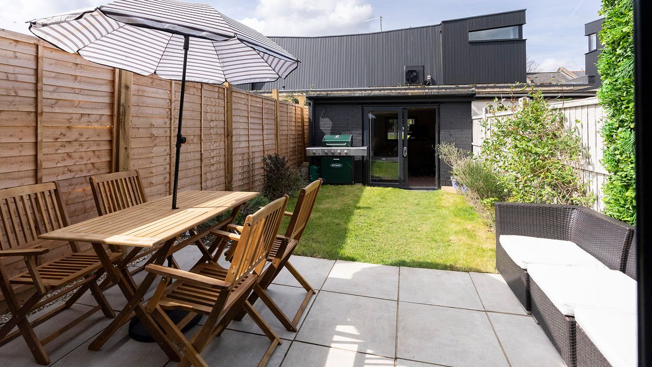 An external shot of backyard with garden outbuilding, wooden garden table and chairs and parasol with striped decor