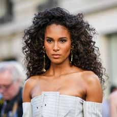 Cindy Bruna wears gold long pendant earrings, a white gray with small black striped print pattern shoulder-off / large belted / long sleeves corset jacket from Jean-Paul Gaultier, matching white gray with small black striped print pattern wide legs suit pants, outside Jean-Paul Gaultier, during Paris Fashion Week - Haute Couture Fall Winter 2022 2023, on July 06, 2022 in Paris, France.