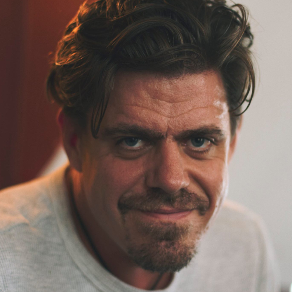 male with dark swept back wavy hair, facial hair wearing light round necked top