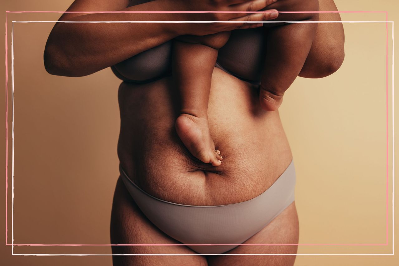 Midsection of mother carrying child while standing against brown background. Postpartum belly with stretch marks.