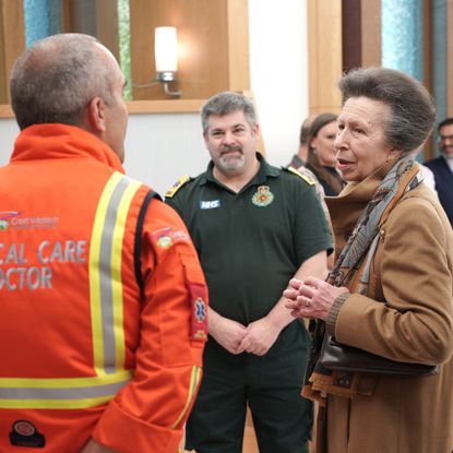 Princess Anne wearing a brown coat standing and talking with doctors at a hospital including a man with an orange coat reading "critical care doctor"