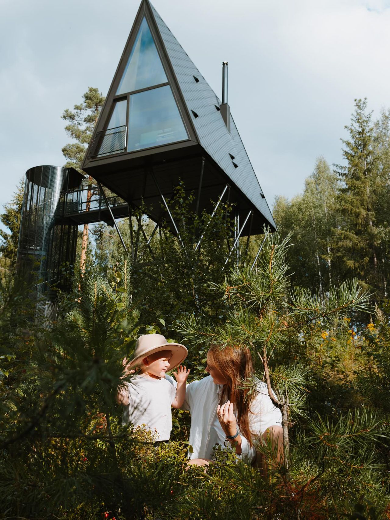 A mum and her young daughter play in a wood underneath a pointy design cabin,