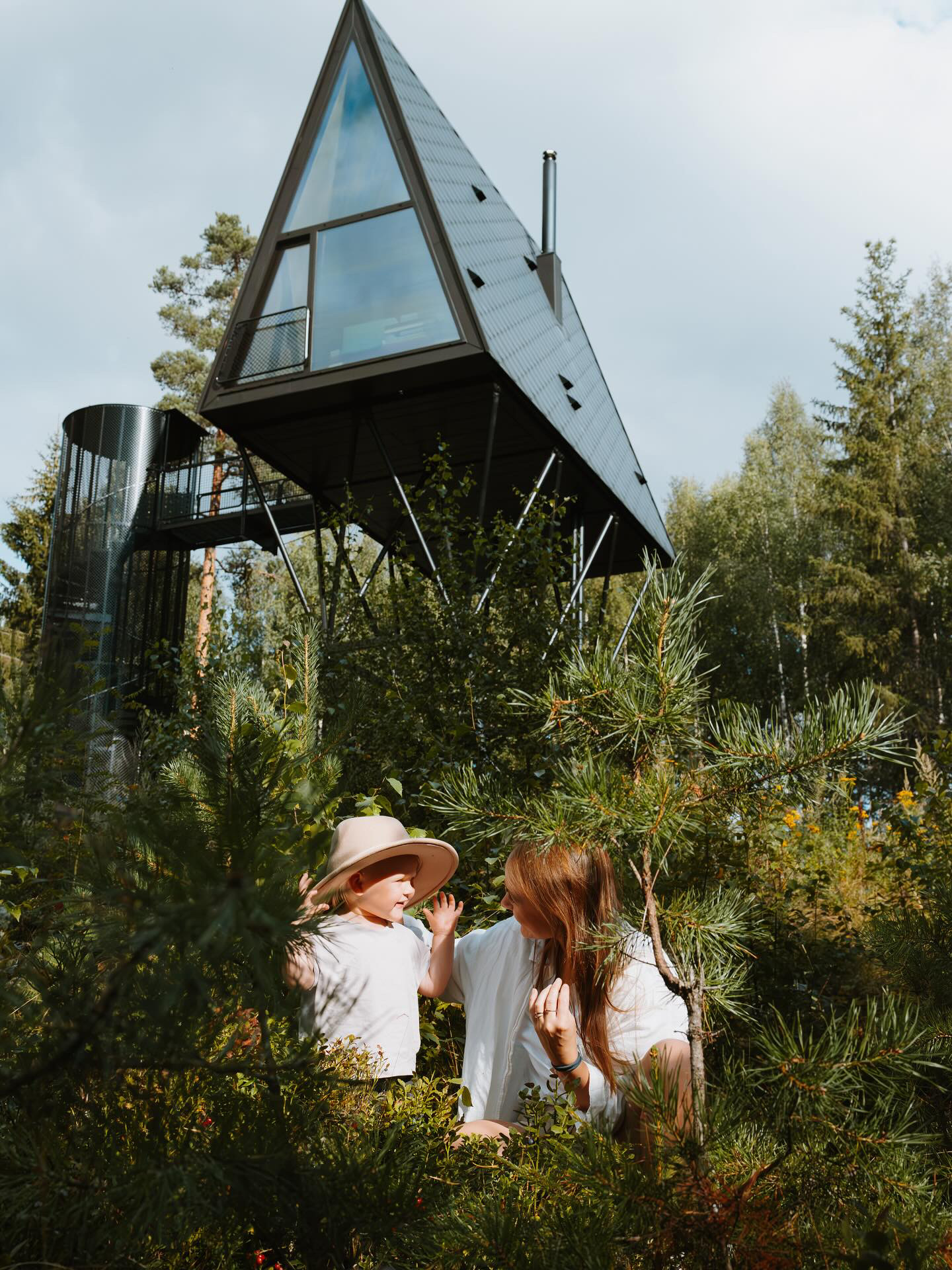 A mum and her young daughter play in a wood underneath a pointy design cabin, 