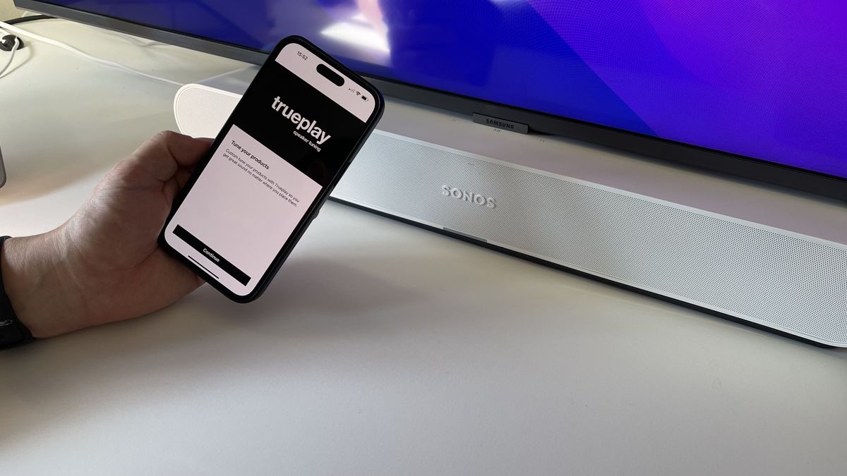 A white Sonos Ray on a white desk, with a phone held in front displaying the Trueplay feature of the Sonos app
