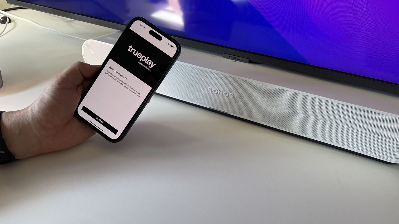 A white Sonos Ray on a white desk, with a phone held in front displaying the Trueplay feature of the Sonos app