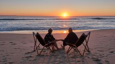 An older couple sit on the beach as the sun sets over the water.