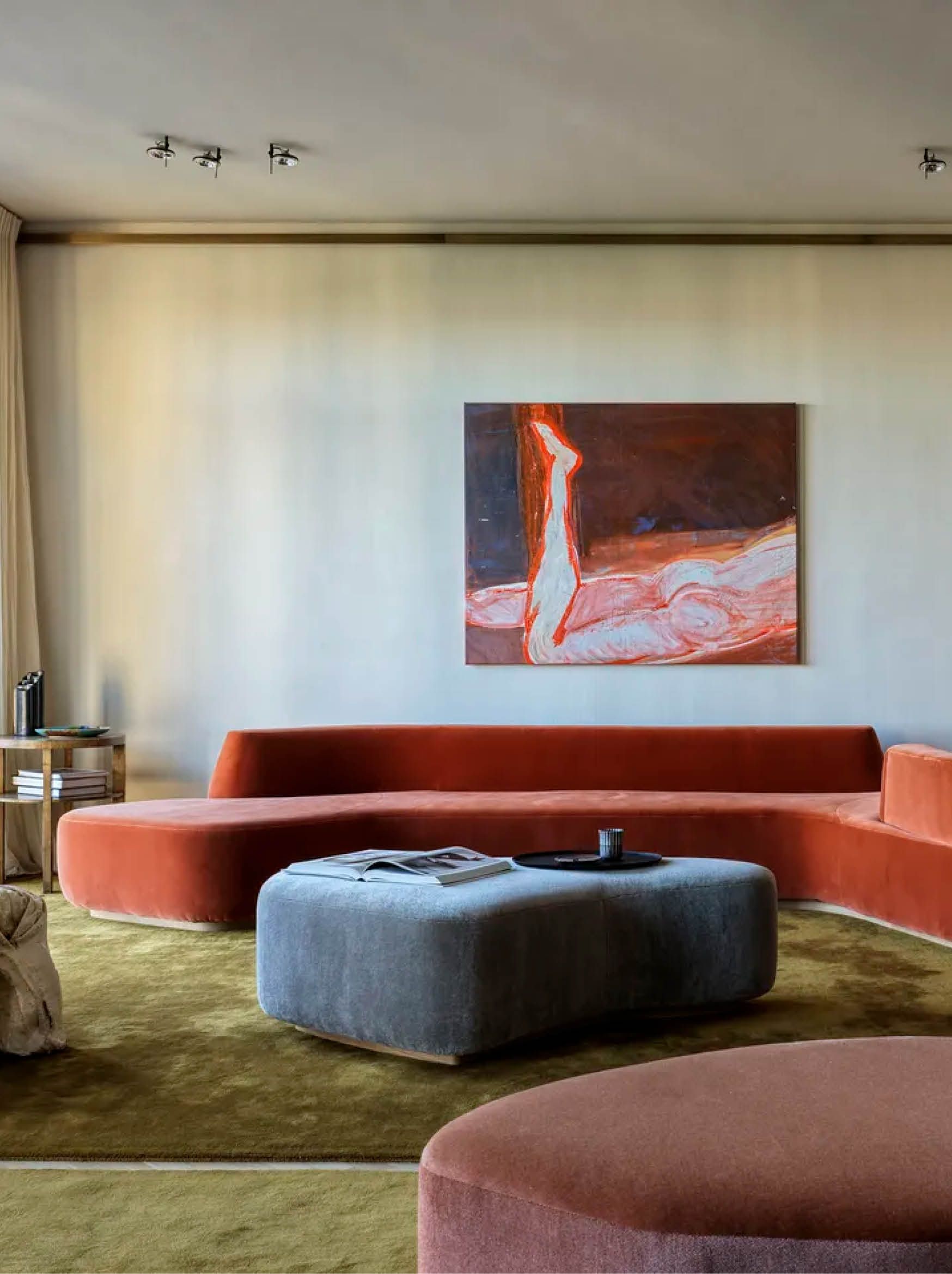 A modern living room with a sculptural red sofa and green carpet, a piece of art on the wall and a gray-blue ottoman