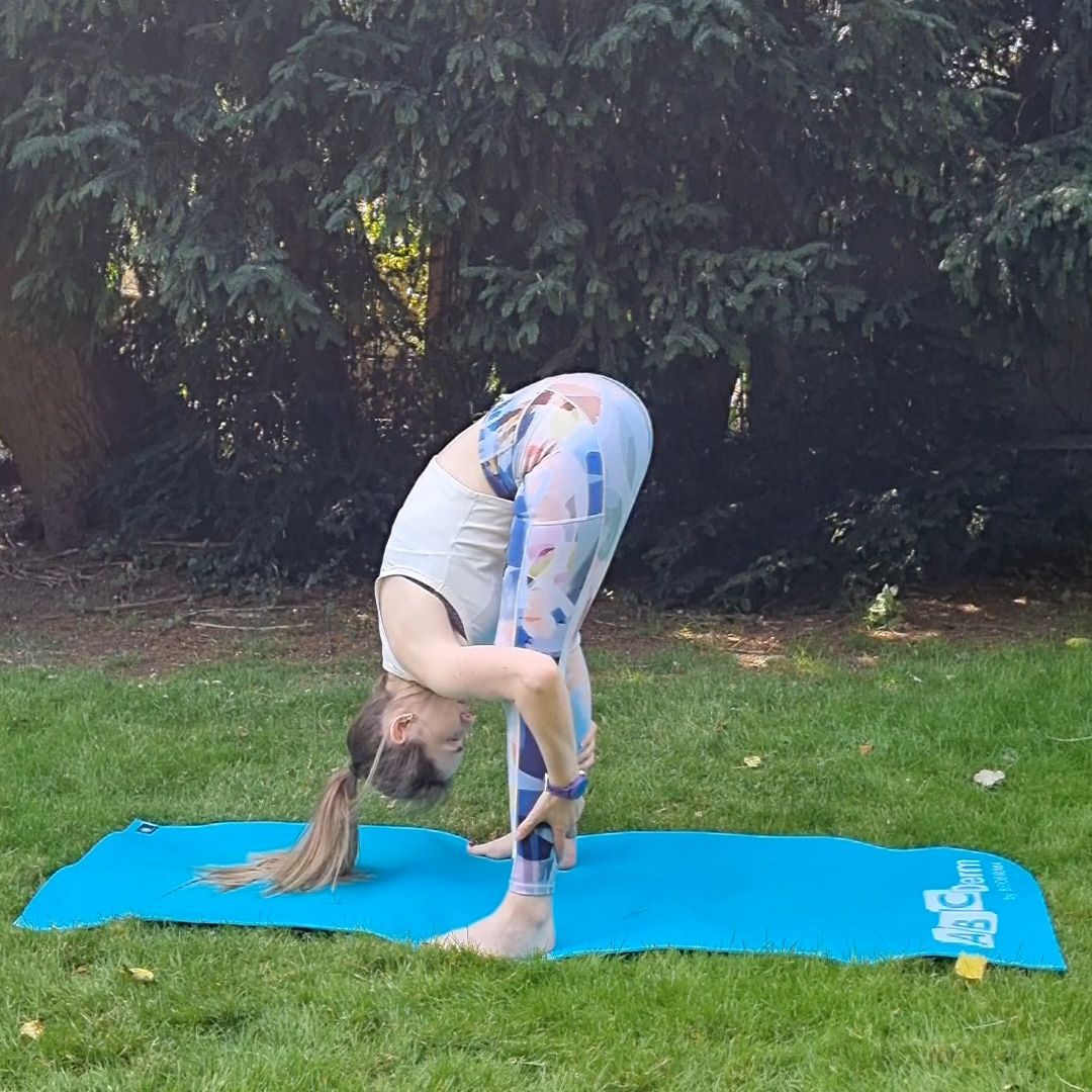 Fitness editor Ruth Gaukrodger performs a deep squat to forward fold. She is in sportswear and exercising on a yoga mat in a green, leafy, outdoor setting. In this picture, her legs are straight and she is hinging at the hips, her torso folded over her legs and arms wrapped around her calves.