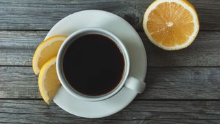 White cup of black coffee with lemon wedges around the edge, sitting on dark wooden table