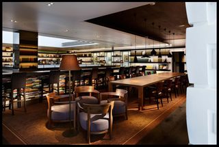 Interior view of the Anjin lounge bar featuring wood flooring, a brown rug, black pendant lights, spotlights, a pillar with a reflective surface, wall-mounted shelving filled with items, framed wall art, a floor lamp, a wooden table, chairs and a bar counter made up of stacked books