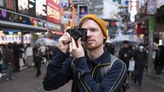 Personne tenant l'appareil photo Fujifilm X100VI à hauteur de l'œil sur fond de ville animée de Tokyo. 