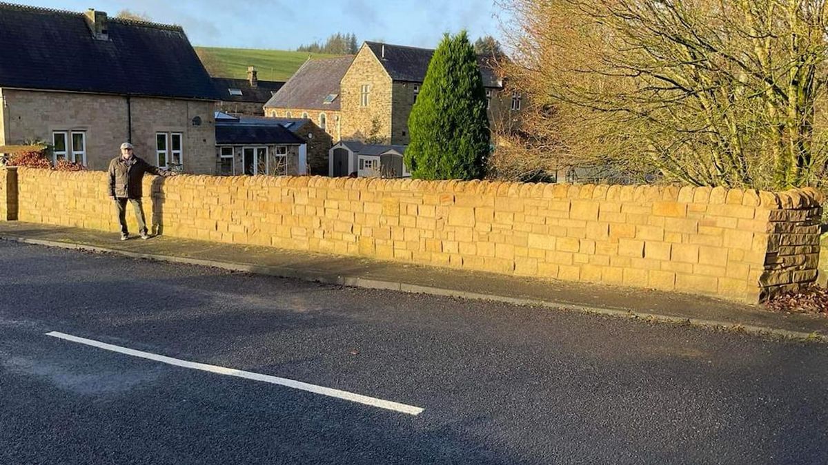 A man standing next to a long sandstone garden wall next to a road