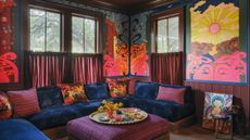 A vibrant, colorful living room corner with a built-in sofa with plush blue velvet seating and colorful scatter cushions, a purple striped pouf in the center with a round tray with plates and decor