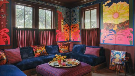 A vibrant, colorful living room corner with a built-in sofa with plush blue velvet seating and colorful scatter cushions, a purple striped pouf in the center with a round tray with plates and decor
