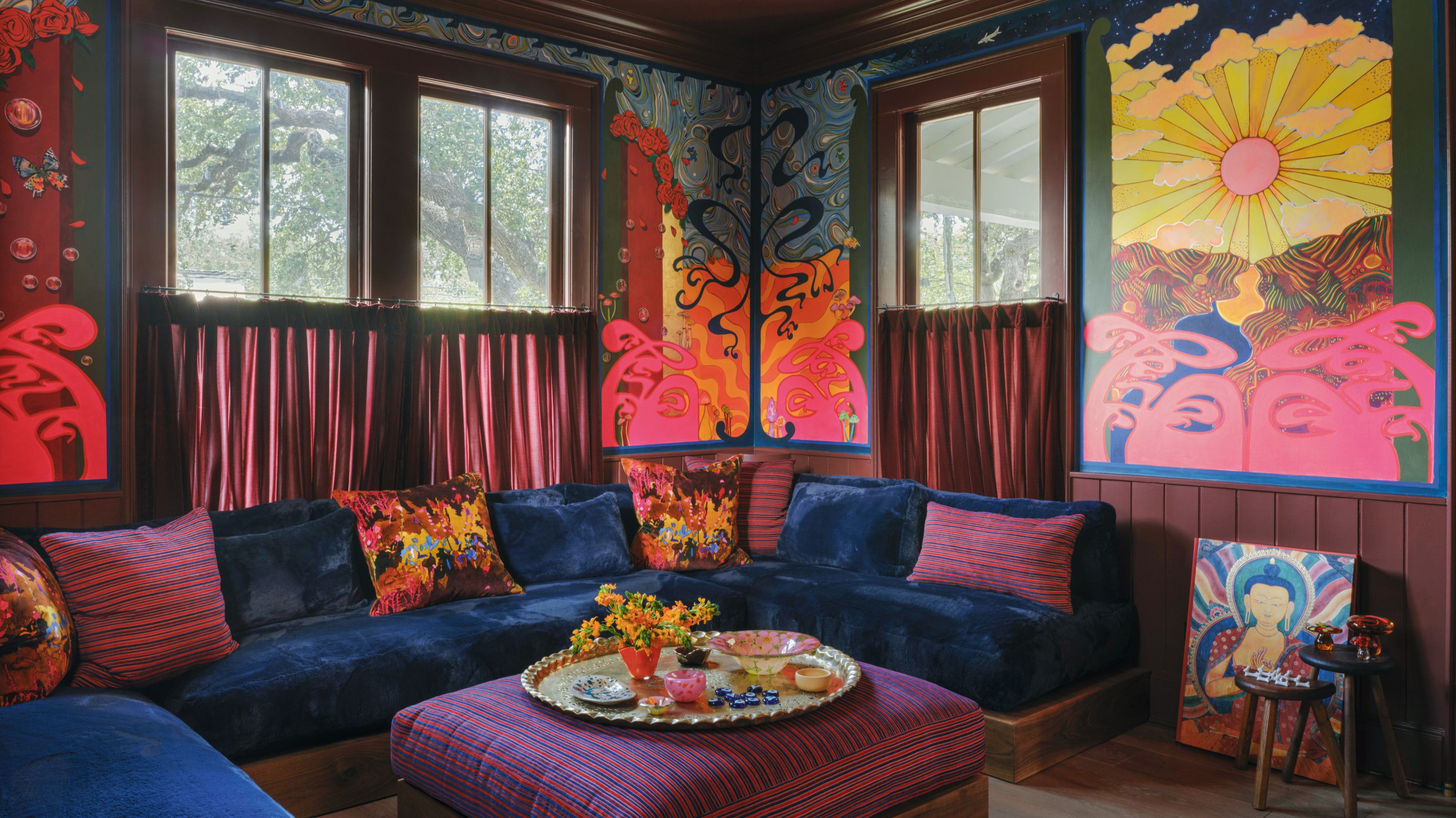 A vibrant, colorful living room corner with a built-in sofa with plush blue velvet seating and colorful scatter cushions, a purple striped pouf in the center with a round tray with plates and decor