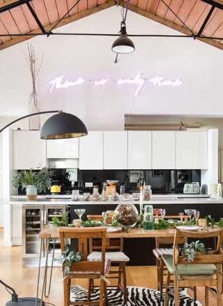 A white kitchen with black gloss backsplash, pink neon slogan light fixture, wooden dining set and faux Zebra rug