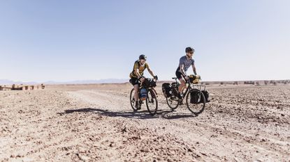 best gravel bike: Two smiling caucasian cyclists ride a desert dirt road in Morocco 