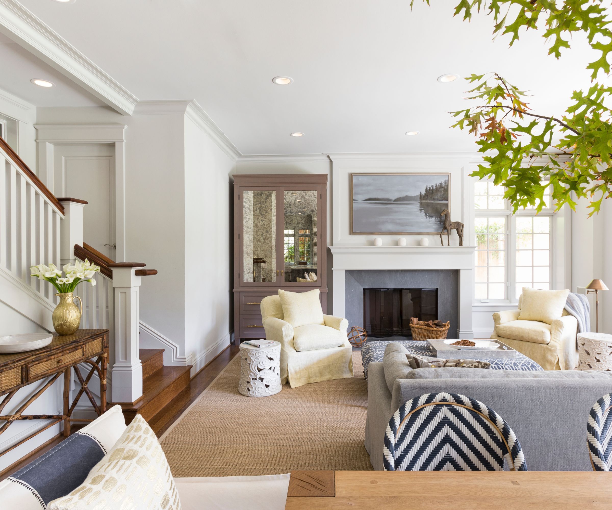Transitional style living room with Gray Owl by Benjamin Moore painted walls, a fireplace with a landscape painting above it, linen armchairs, a jute rug, a wooden staircase and a mirrored armoire.