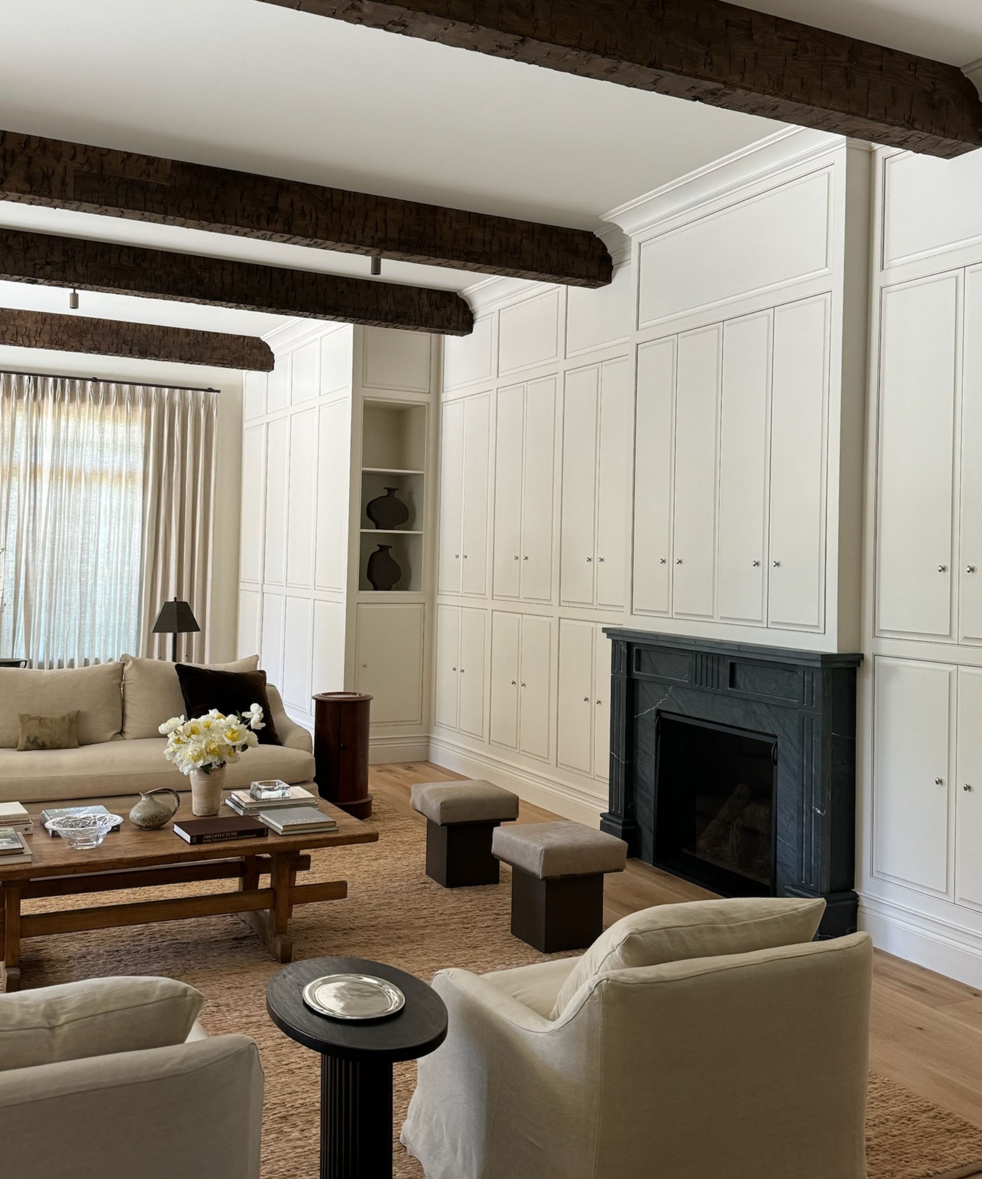 White living room with bespoke cabinetry and black marble fireplace to the right, seating around a wooden coffee table to the left, window with sheer curatins in background