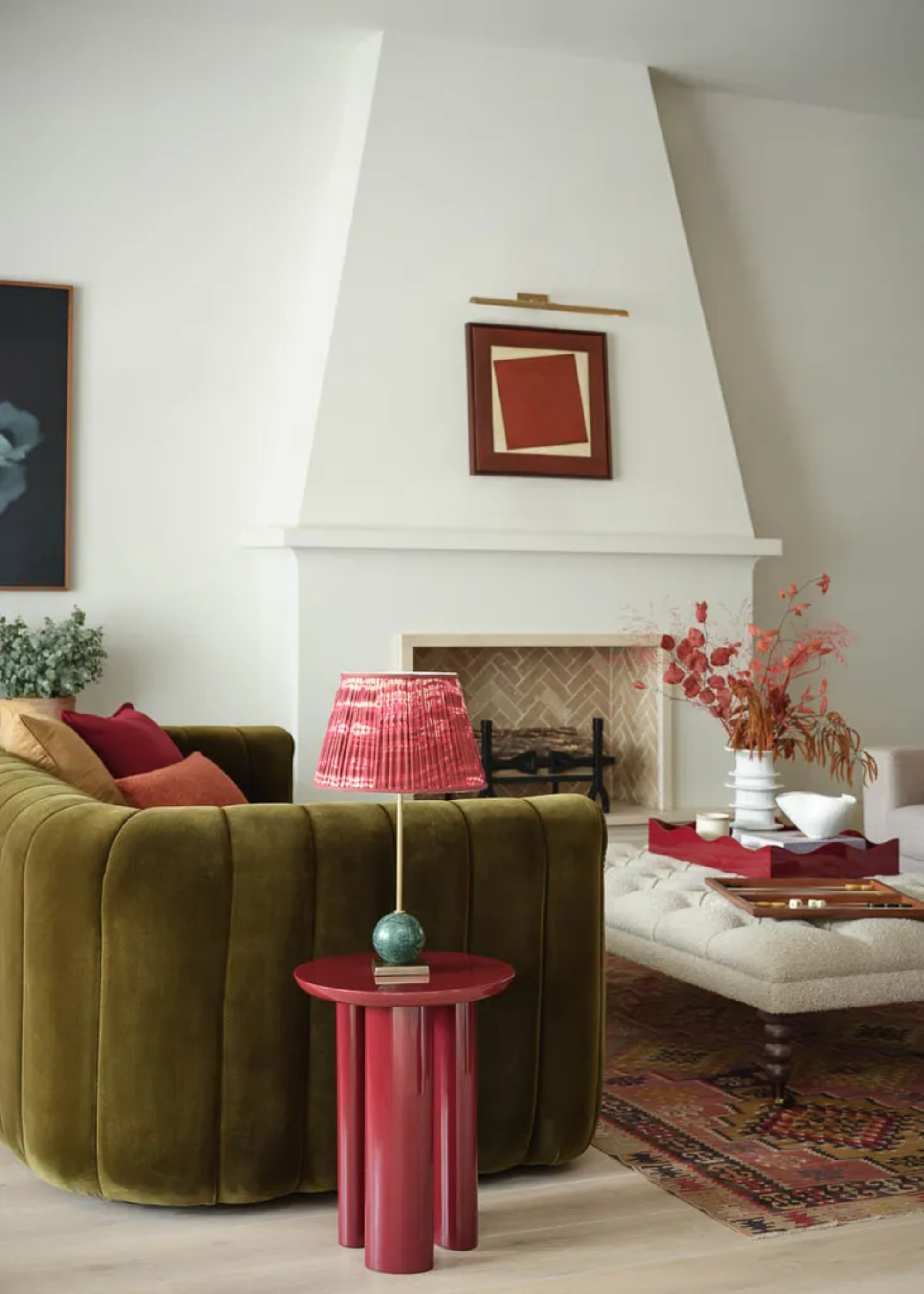 A white living room with an olive green couch, a Persian rug, an upholstered coffee table, a maroon side table and a table lamp