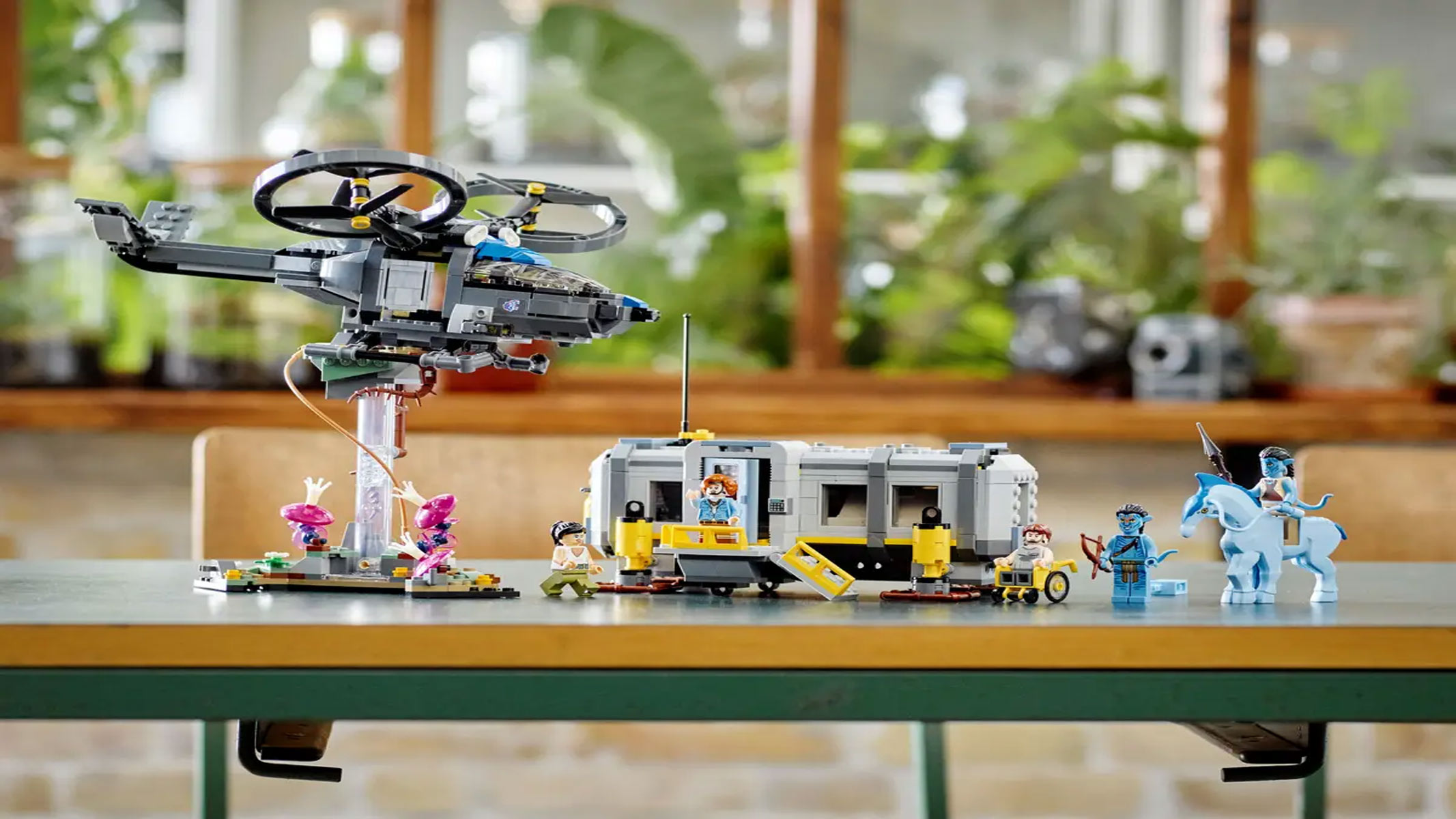 A brick-built helicopter is displayed on a stand next to a mini building and a few minifigures, all on a table with plants in the background.
