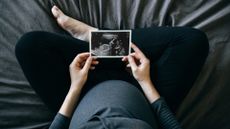 A top down shot of a pregnant person wearing a blue shirt and black pants sitting cross-legged on a blue bed, holding an ultrasound in front of them. 