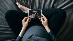 A top down shot of a pregnant person wearing a blue shirt and black pants sitting cross-legged on a blue bed, holding an ultrasound in front of them. 