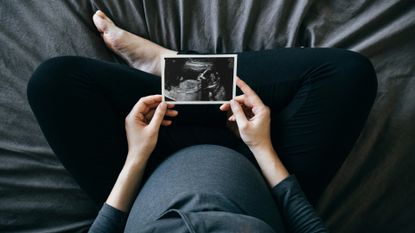 A top down shot of a pregnant person wearing a blue shirt and black pants sitting cross-legged on a blue bed, holding an ultrasound in front of them.