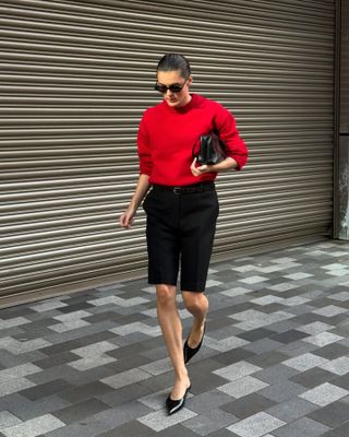 Die Mode-Influencerin Valeria @sobalera spaziert über den Londoner Bürgersteig und trägt einen ordentlich gekämmten Dutt, eine ovale Sonnenbrille, einen roten Pullover mit Rundhalsausschnitt, eine schwarze Tasche, die als Clutch getragen wird, schwarze Bermudashorts mit Gürtel und schwarze, spitze Sandaletten mit Kitten-Heels.