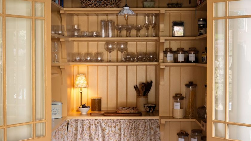 Charming cottage-style pantry with soft wood shelving, glassware displays, labelled jars, warm lighting, and a fabric-curtained counter creating a cosy, lived-in feel.