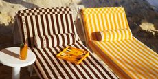 Two stripy sun loungers, one brown and cream, one yellow and cream, are placed on a sandy beach out in natural light next to a round coffee table topped with an orangey cocktail.