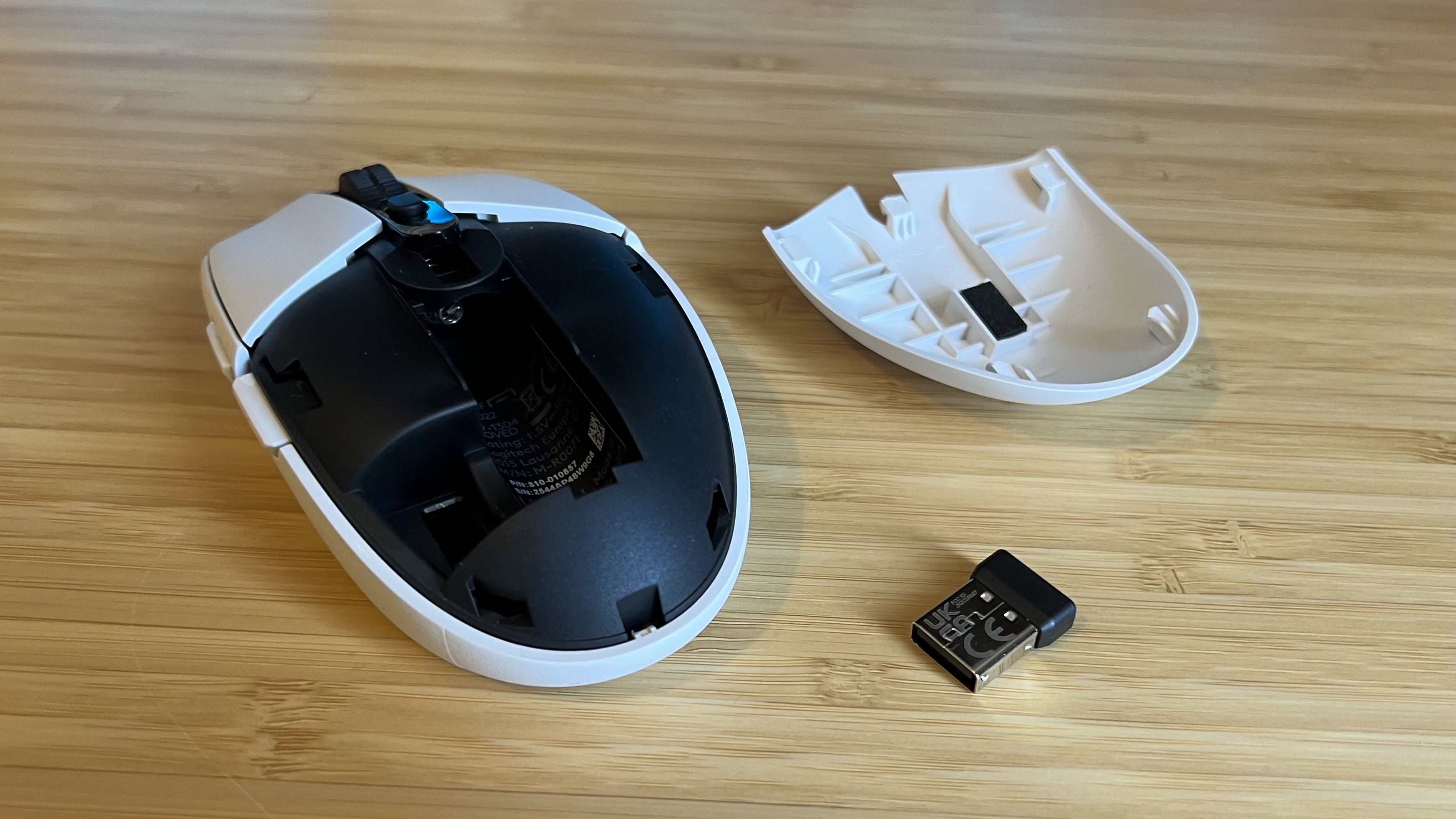 Logitech G305 Lightspeed gaming mouse with top cover removed showing battery compartment and 2.4GHz dongle on a wooden desk