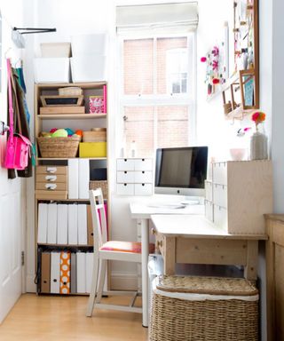 A white home office with brown shelves with files on them, a white desk with a silver computer screen and chair next to it and window above it, a wooden table with a storage box on it, and a wicker bin