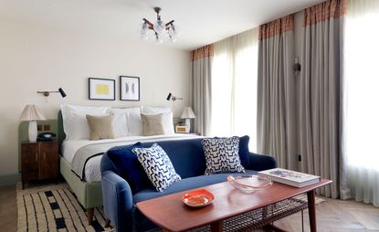 A room in the Redchurch Townhouse hotel. The large, pale green bed is covered in white linen with a deep blue couch and a wooden table in front of it. The walls are painted in light cream color and there are two large windows covered with see-through curtains.