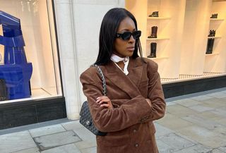 British influencer @georgia_3.0 wearing a brown suede blazer, white jeans, Chanel Classic Flap Bag, and black sunglasses.