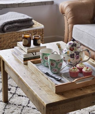 A traditional tea pot and mugs containing English breakfast tea on wooden table with Berber rug or carpet underneath in living room with tan sofa decor