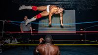 A wrestler in white trunks and red boots leaps mid-air inside a wrestling ring. Another wrestler stands below, surprised. A referee observes the scene
