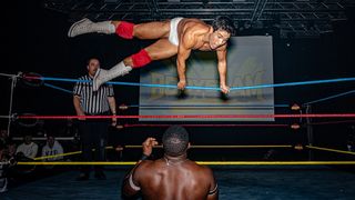 A wrestler in white trunks and red boots leaps mid-air inside a wrestling ring. Another wrestler stands below, surprised. A referee observes the scene