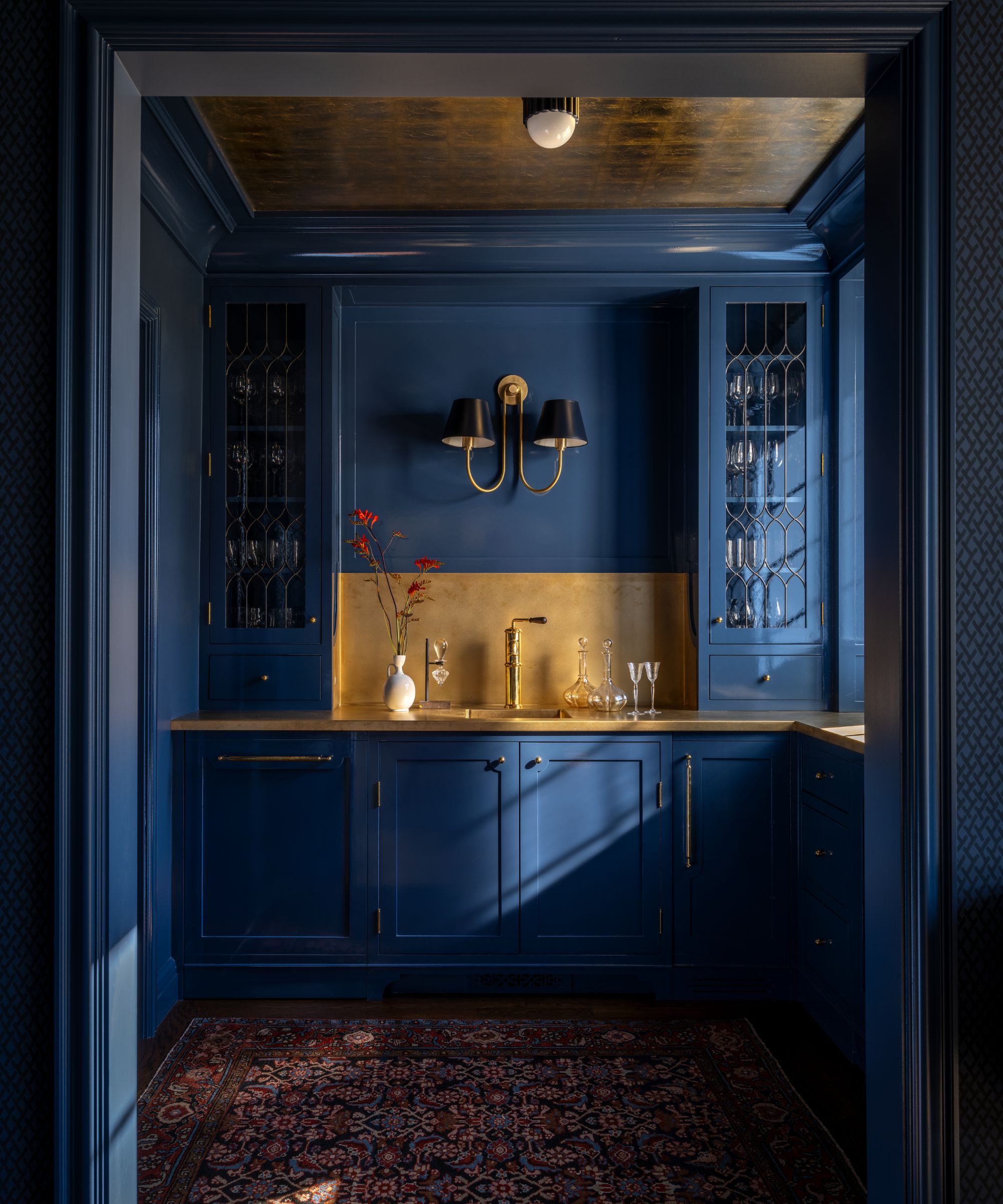 A home bar room with dark blue walls and cabinets, brass counters, and a red patterned rug on the ground.