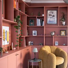 A living room with built-in bookshelves and cabinets painted in a dusky rose pink and filled with framed pictures, plants and books and decorative objects