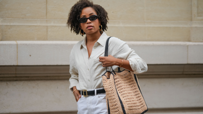 Ellie Delphine wears a full Mango look, sunglasses, golden earrings, a white linen shirt, a black leather belt with golden buckle, golden bracelets, a white ripped denim shorts, a large brown raffia bag, during a street style fashion photo session, on May 08, 2024 in Paris, France. 