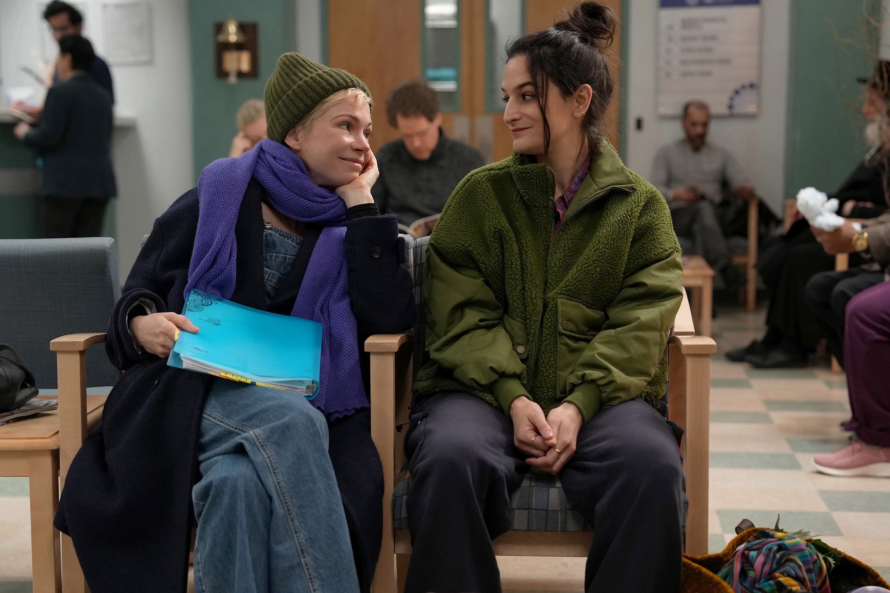 michelle williams as molly wearing a hat and scarf and jenny slate as nikki wearing a jacket sitting in a doctor's office lobby in dying for sex