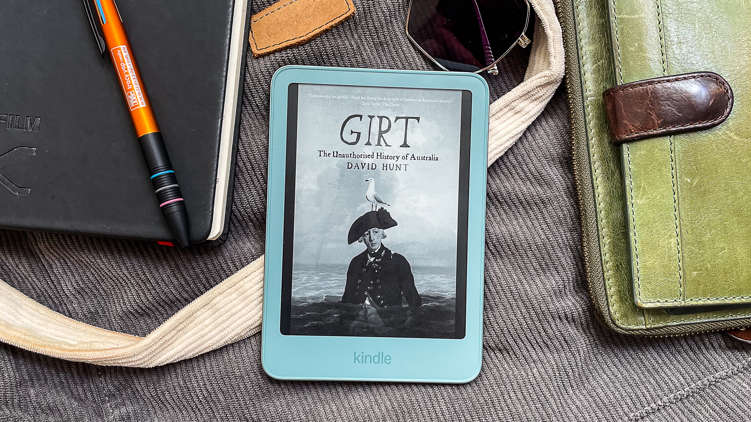 A book cover displayed on the Amazon Kindle 2026 while it lies on a cloth bag and surrounded by handbag contents