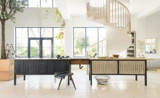 kitchen by deVOL and Sebastian Cox, neutral room, large black frame windows looking on into the garden, neutral gloss flooring, tree planter in the left corner, spiral staircase, stepladders, hanging plants, radiator, black and wooden centre piece worktops, wooden stool with dark apron on, basket on the floor, wooden drawer unit to the left