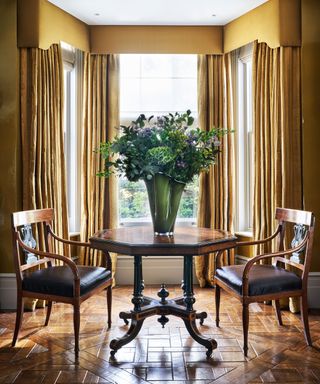 Traditional dining room with dining table with two dining chairs with a vase and large flower arrangements, bay window with yellow curtains and pelmet.