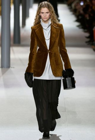 how to wear velvet is shown in a photo from Louis Vuitton's fall 2024 runway show with a model wearing a brown velvet blazer over a white funnel-neck blouse with sheer pants and furry gloves