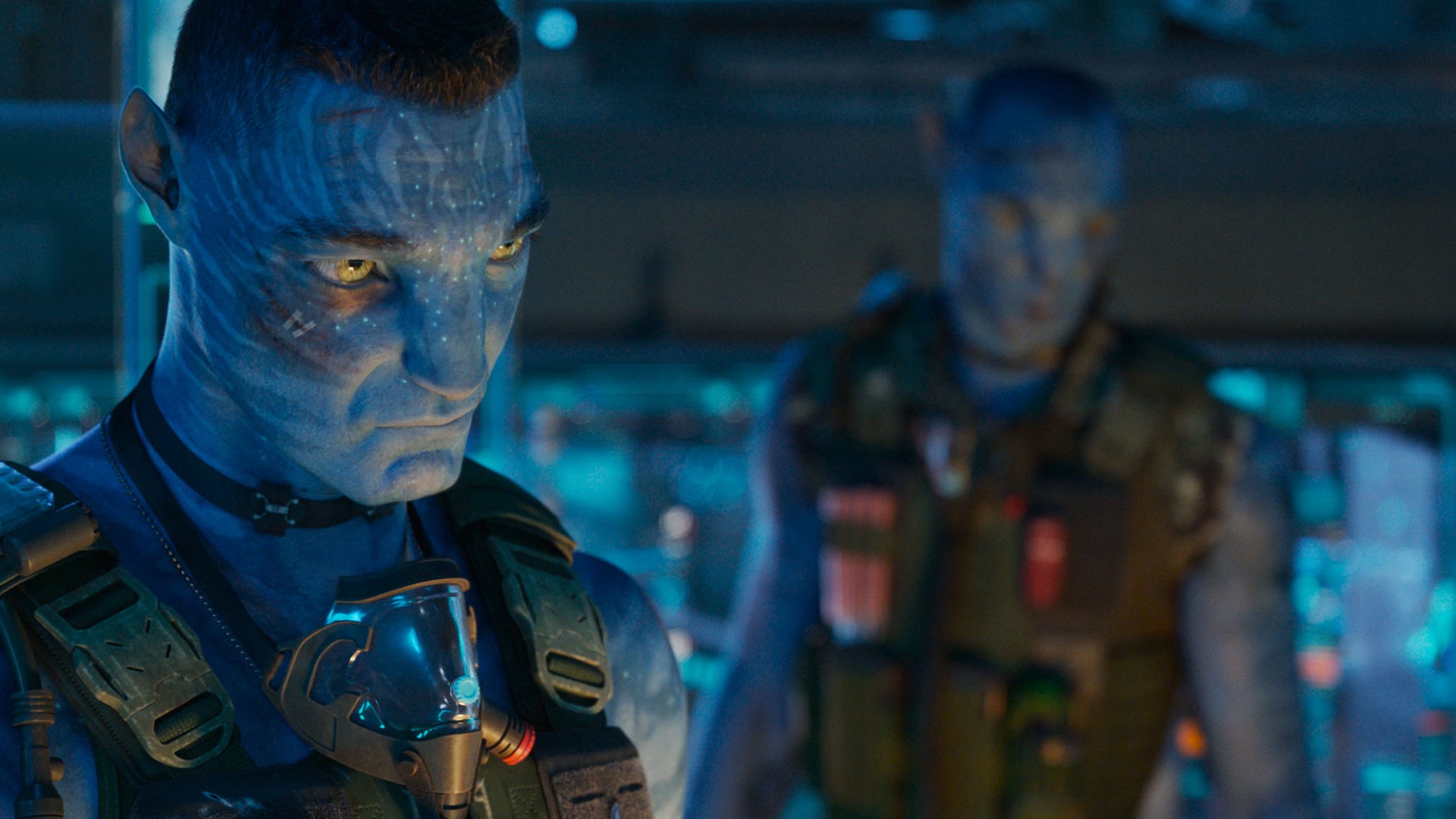 Still from the movie Avatar: Fire and Ash. Two blue-skinned, yellow-eyed Na-vi male are standing in a room with a lot of hi-tech screens. They are both wearing a protective vest and each have a breathing mask hanging around their neck.