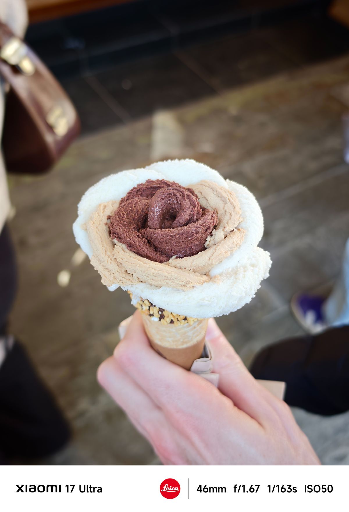 Hand holding an ice cream cone topped with chocolate and vanilla scoops arranged like a flower, shallow depth of field background.