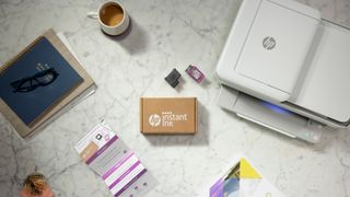An instant ink pack sits by an HP printer, a coffee and some spectacles