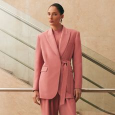 Model wearing a peachy pink suit with side tie detail wearing large sliver drop earrings sold at Karen Millen in front of a minimal staircase for Karen Millen Discount Codes